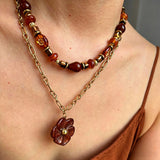 This vintage‑inspired Meidelat layered necklace features warm amber‑toned beads paired with polished gold‑toned metal chains. A carved rich brown flower pendant hangs on the lower paperclip chain, while the upper strand mixes varied-shaped resin beads for textured depth. Perfect for creating cozy boho‑chic layered neck stacking looks.