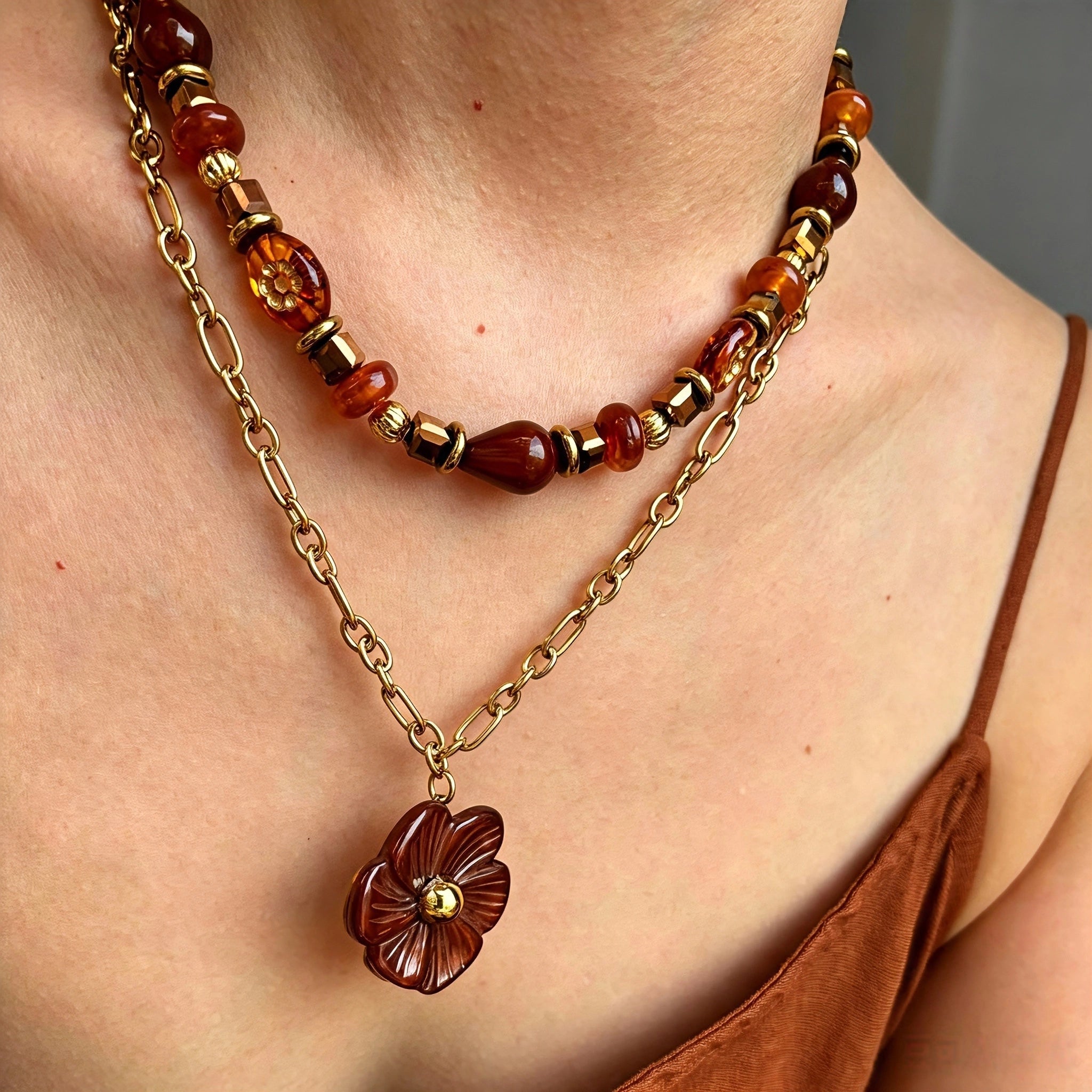 This vintage‑inspired Meidelat layered necklace features warm amber‑toned beads paired with polished gold‑toned metal chains. A carved rich brown flower pendant hangs on the lower paperclip chain, while the upper strand mixes varied-shaped resin beads for textured depth. Perfect for creating cozy boho‑chic layered neck stacking looks.