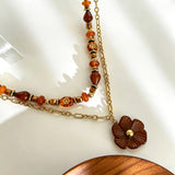 This vintage‑inspired Meidelat layered necklace features warm amber‑toned beads paired with polished gold‑toned metal chains. A carved rich brown flower pendant hangs on the lower paperclip chain, while the upper strand mixes varied-shaped resin beads for textured depth. Perfect for creating cozy boho‑chic layered neck stacking looks.