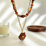 This vintage‑inspired Meidelat layered necklace features warm amber‑toned beads paired with polished gold‑toned metal chains. A carved rich brown flower pendant hangs on the lower paperclip chain, while the upper strand mixes varied-shaped resin beads for textured depth. Perfect for creating cozy boho‑chic layered neck stacking looks.