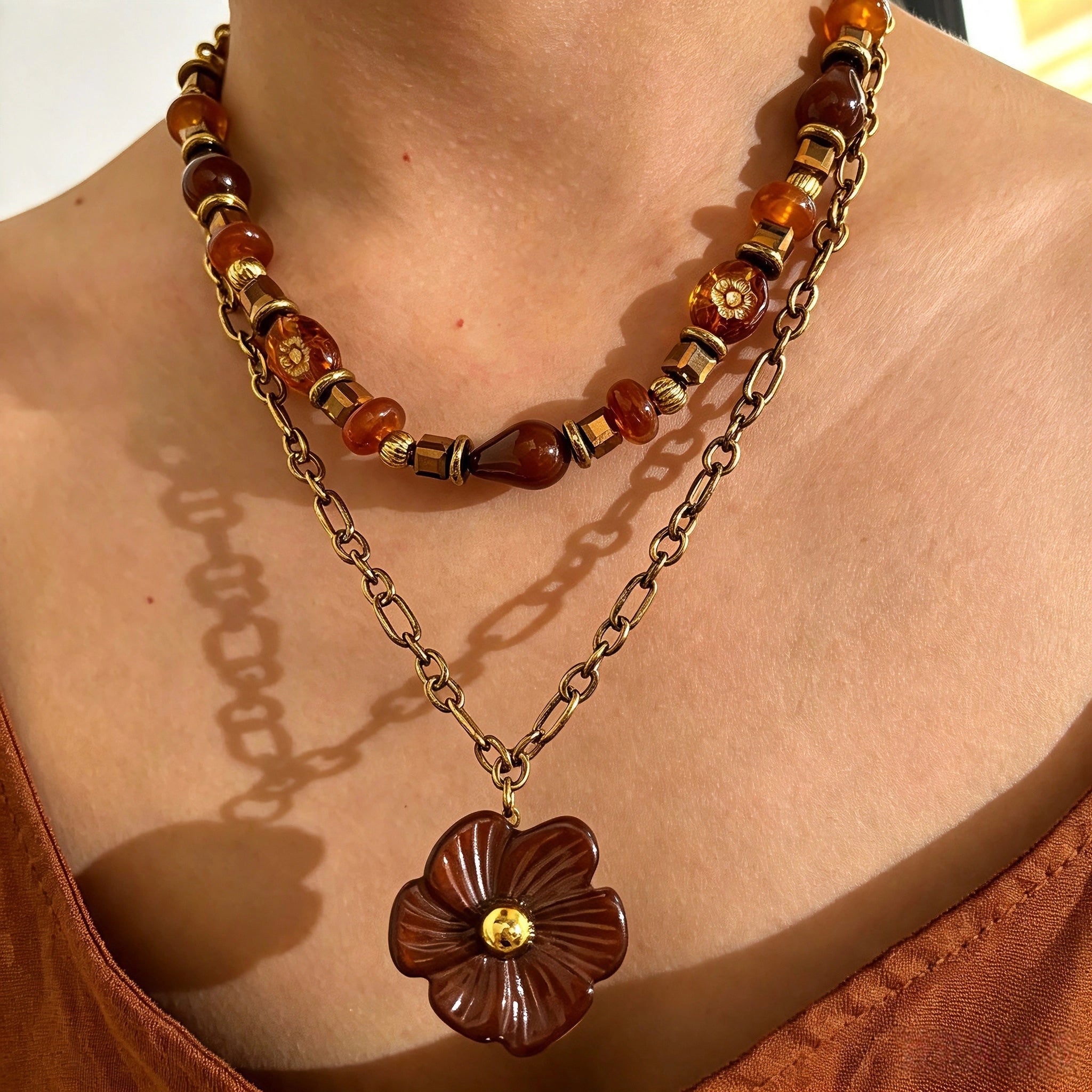 This vintage‑inspired Meidelat layered necklace features warm amber‑toned beads paired with polished gold‑toned metal chains. A carved rich brown flower pendant hangs on the lower paperclip chain, while the upper strand mixes varied-shaped resin beads for textured depth. Perfect for creating cozy boho‑chic layered neck stacking looks.