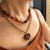 This vintage‑inspired Meidelat layered necklace features warm amber‑toned beads paired with polished gold‑toned metal chains. A carved rich brown flower pendant hangs on the lower paperclip chain, while the upper strand mixes varied-shaped resin beads for textured depth. Perfect for creating cozy boho‑chic layered neck stacking looks.