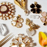 These four-leaf clover brooches come in two elegant versions: one with glossy black onyx petals centered by a delicate pearl, and the other with milky white crystal petals accented by a deep red gemstone at its core. Both are framed in polished gold-toned metal, exuding a classic, vintage-inspired charm.