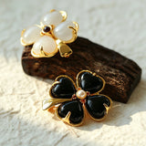 These four-leaf clover brooches come in two elegant versions: one with glossy black onyx petals centered by a delicate pearl, and the other with milky white crystal petals accented by a deep red gemstone at its core. Both are framed in polished gold-toned metal, exuding a classic, vintage-inspired charm.