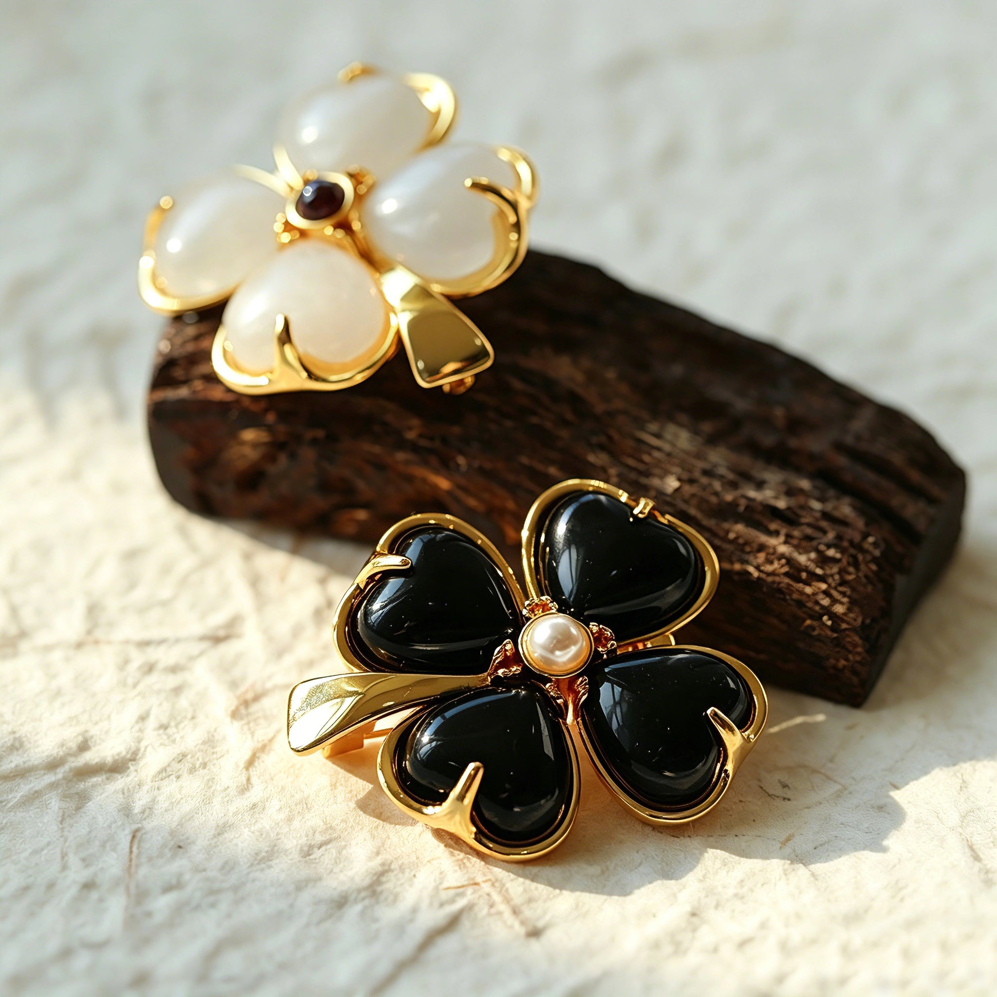 These four-leaf clover brooches come in two elegant versions: one with glossy black onyx petals centered by a delicate pearl, and the other with milky white crystal petals accented by a deep red gemstone at its core. Both are framed in polished gold-toned metal, exuding a classic, vintage-inspired charm.