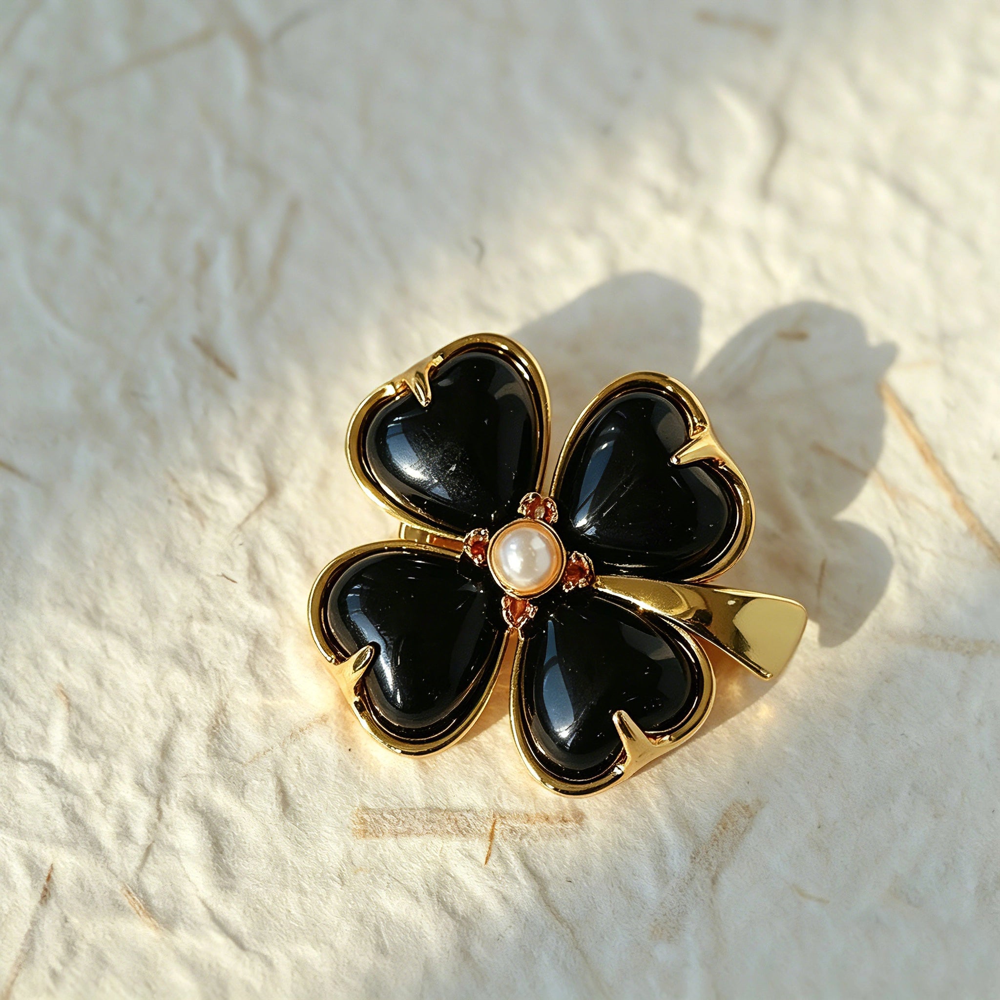 These four-leaf clover brooches come in two elegant versions: one with glossy black onyx petals centered by a delicate pearl, and the other with milky white crystal petals accented by a deep red gemstone at its core. Both are framed in polished gold-toned metal, exuding a classic, vintage-inspired charm.