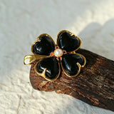 These four-leaf clover brooches come in two elegant versions: one with glossy black onyx petals centered by a delicate pearl, and the other with milky white crystal petals accented by a deep red gemstone at its core. Both are framed in polished gold-toned metal, exuding a classic, vintage-inspired charm.