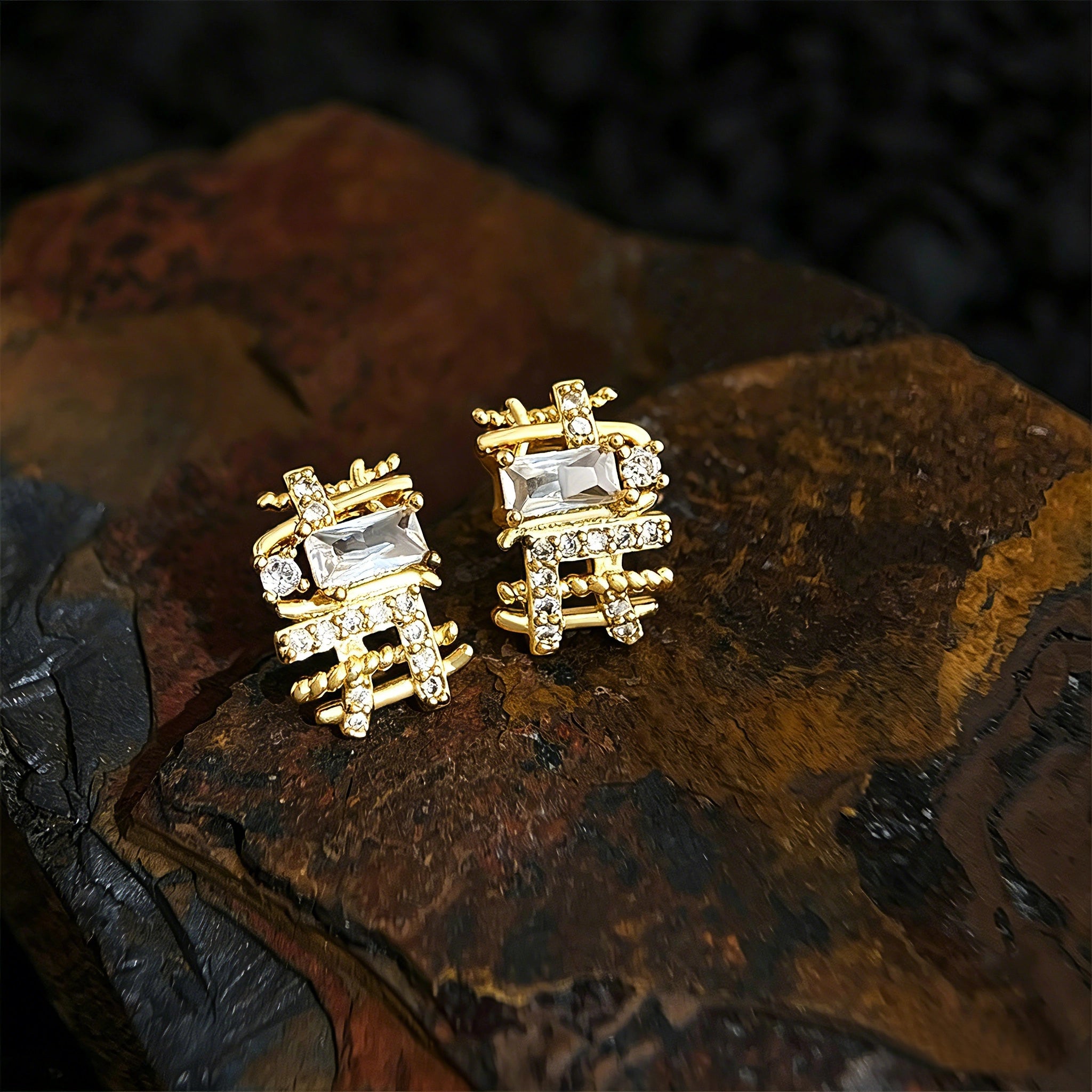 These 18k real gold-plated stud earrings feature an intricate geometric woven design, crafted with twisted gold tones that create a luxurious, textured look. Adorned with a central rectangular baguette-cut zirconia stone surrounded by round pavé-set zircons, they sparkle brilliantly with a blend of clear cubic zirconia and golden metalwork. The elegant yet bold silhouette combines modern geometry with timeless glamour, making it a striking accessory.