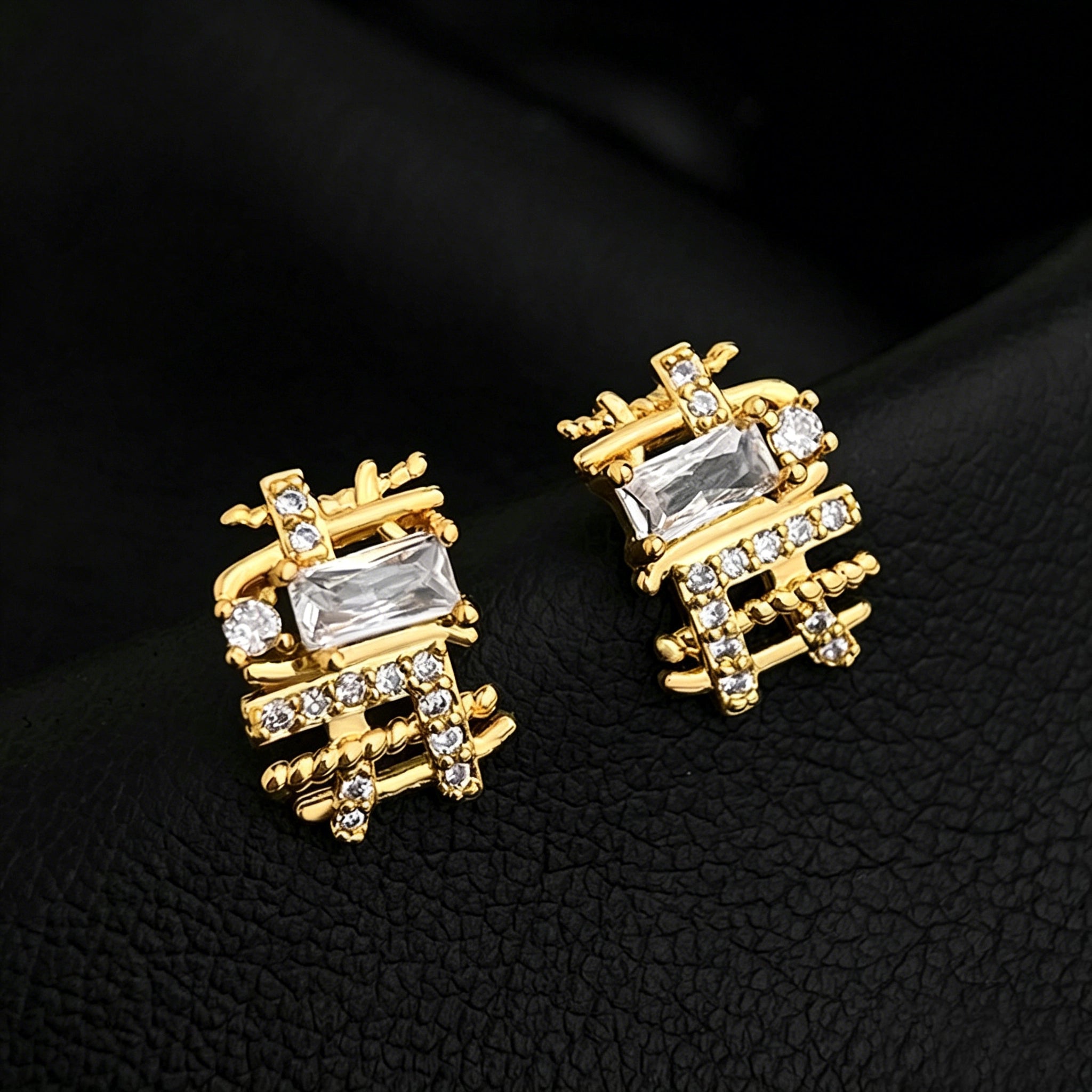 These 18k real gold-plated stud earrings feature an intricate geometric woven design, crafted with twisted gold tones that create a luxurious, textured look. Adorned with a central rectangular baguette-cut zirconia stone surrounded by round pavé-set zircons, they sparkle brilliantly with a blend of clear cubic zirconia and golden metalwork. The elegant yet bold silhouette combines modern geometry with timeless glamour, making it a striking accessory.