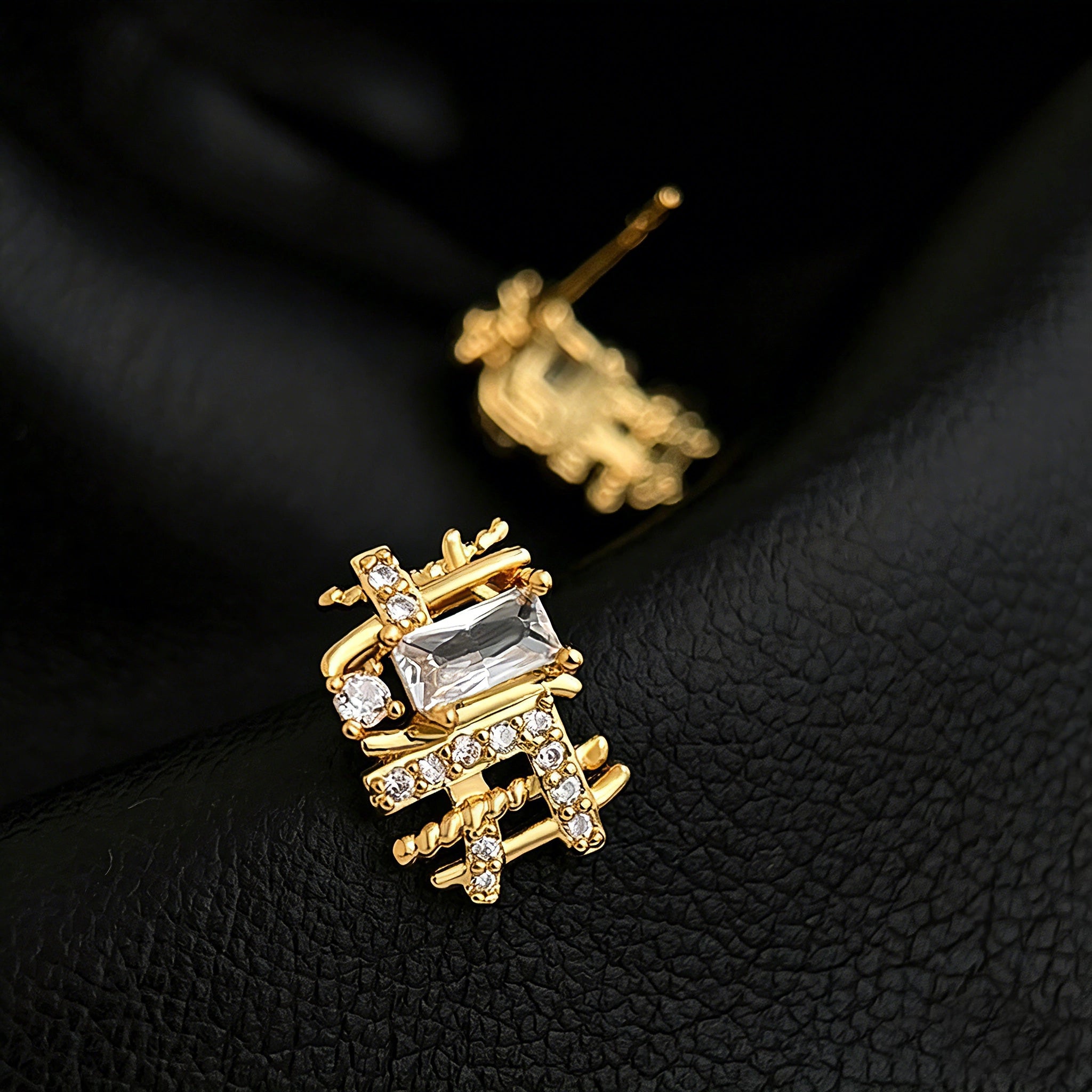 These 18k real gold-plated stud earrings feature an intricate geometric woven design, crafted with twisted gold tones that create a luxurious, textured look. Adorned with a central rectangular baguette-cut zirconia stone surrounded by round pavé-set zircons, they sparkle brilliantly with a blend of clear cubic zirconia and golden metalwork. The elegant yet bold silhouette combines modern geometry with timeless glamour, making it a striking accessory.