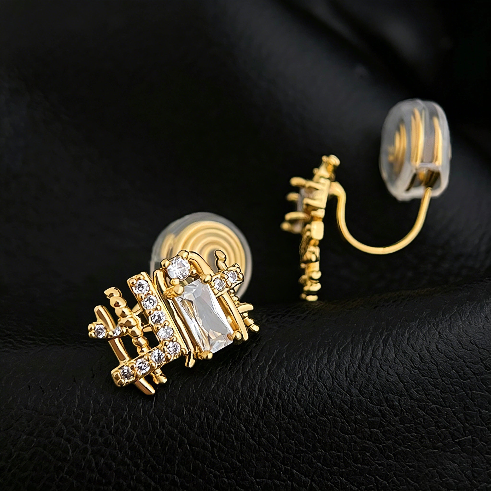 These 18k real gold-plated stud earrings feature an intricate geometric woven design, crafted with twisted gold tones that create a luxurious, textured look. Adorned with a central rectangular baguette-cut zirconia stone surrounded by round pavé-set zircons, they sparkle brilliantly with a blend of clear cubic zirconia and golden metalwork. The elegant yet bold silhouette combines modern geometry with timeless glamour, making it a striking accessory.
