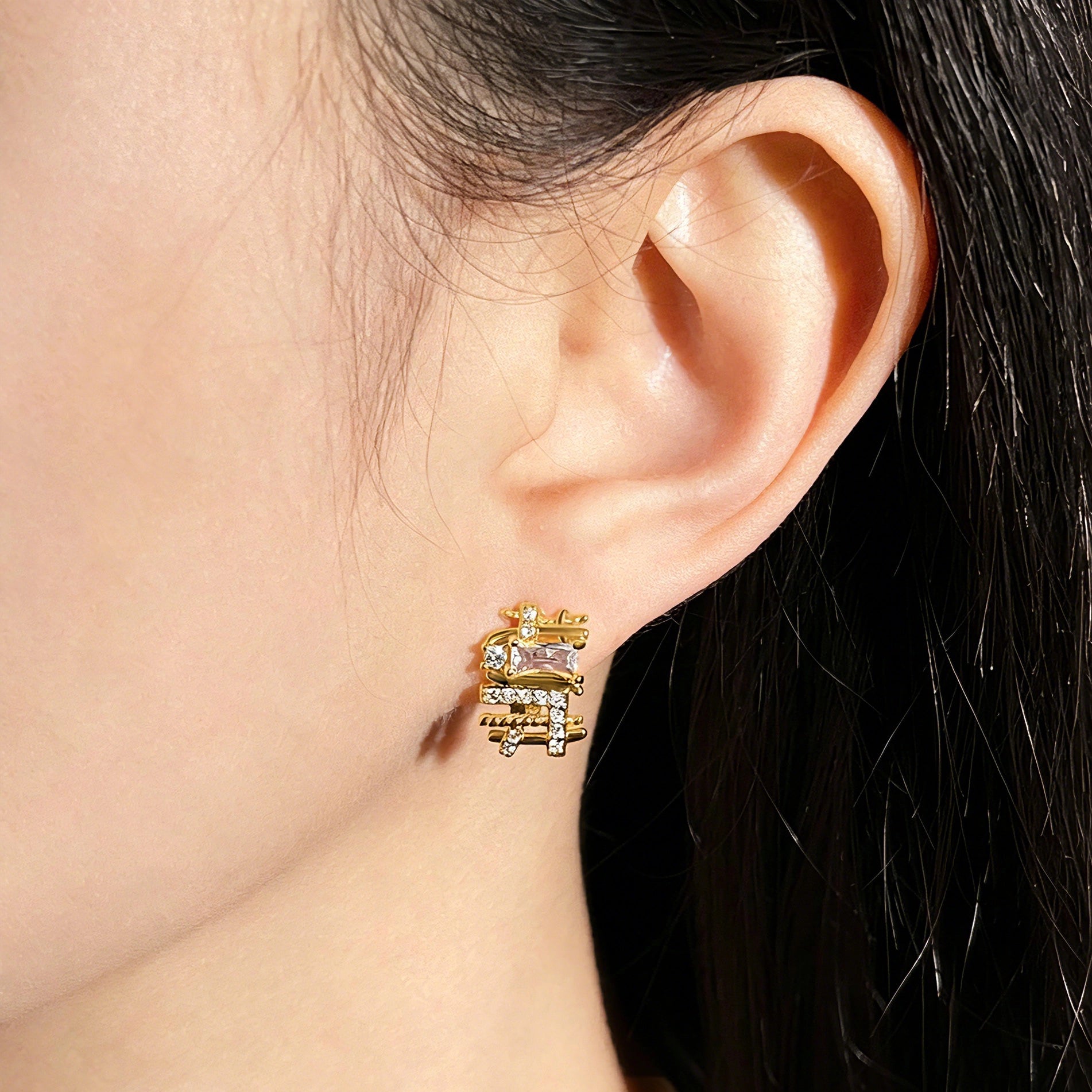 These 18k real gold-plated stud earrings feature an intricate geometric woven design, crafted with twisted gold tones that create a luxurious, textured look. Adorned with a central rectangular baguette-cut zirconia stone surrounded by round pavé-set zircons, they sparkle brilliantly with a blend of clear cubic zirconia and golden metalwork. The elegant yet bold silhouette combines modern geometry with timeless glamour, making it a striking accessory.