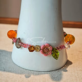 This is a bracelet consist of glass beads. The main color of the bracelet is pink. It's also decorated with orange, yellow pear, grey flower, brown leaves and green leaves spacers.
