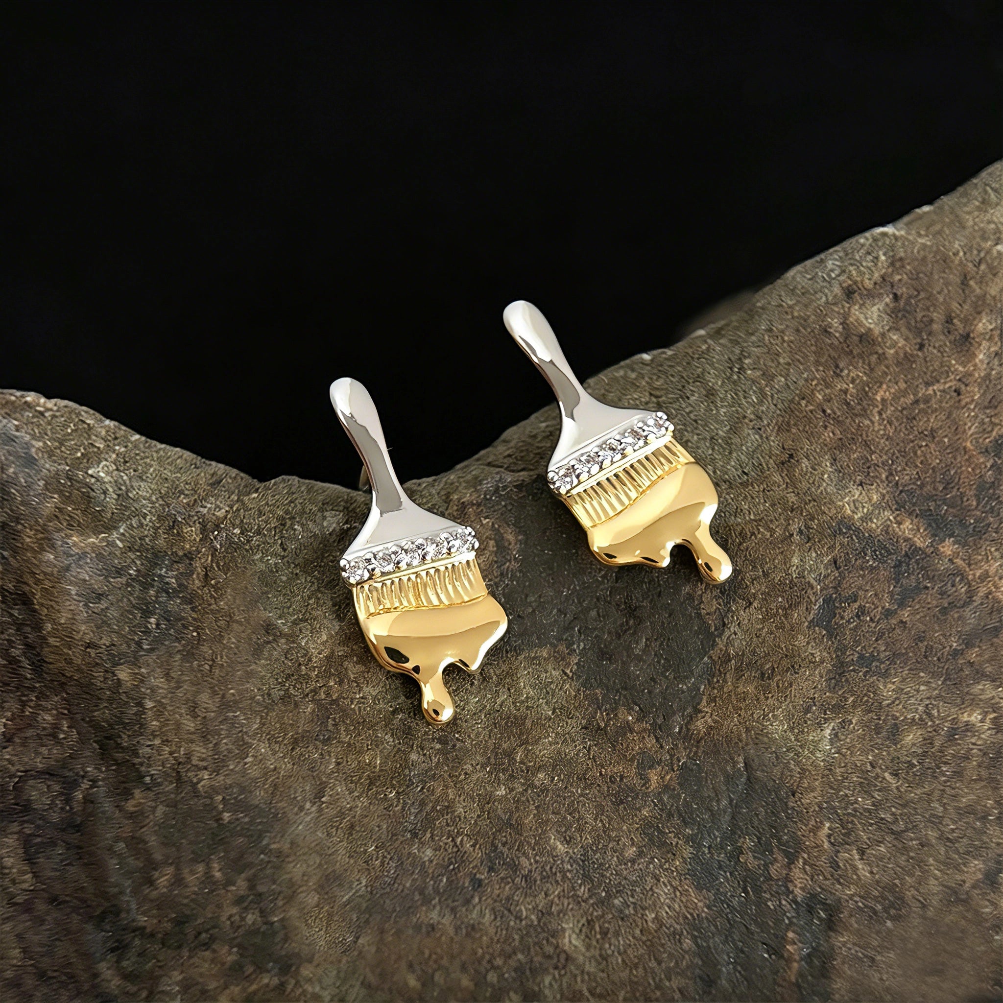 These stud earrings are designed in the shape of a paintbrush, featuring a two-tone finish with a polished silver-toned handle and a gold-toned brush head. The ferrule of the brush is encrusted with sparkling cubic zirconia stones, and the gold-toned bristles are styled to look like dripping, liquid gold paint, creating a vivid, artistic effect. Crafted with a thick 18K real gold plating, they combine a playful, creative design with elegant, eye-catching sparkle.