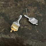 These stud earrings are designed in the shape of a paintbrush, featuring a two-tone finish with a polished silver-toned handle and a gold-toned brush head. The ferrule of the brush is encrusted with sparkling cubic zirconia stones, and the gold-toned bristles are styled to look like dripping, liquid gold paint, creating a vivid, artistic effect. Crafted with a thick 18K real gold plating, they combine a playful, creative design with elegant, eye-catching sparkle.