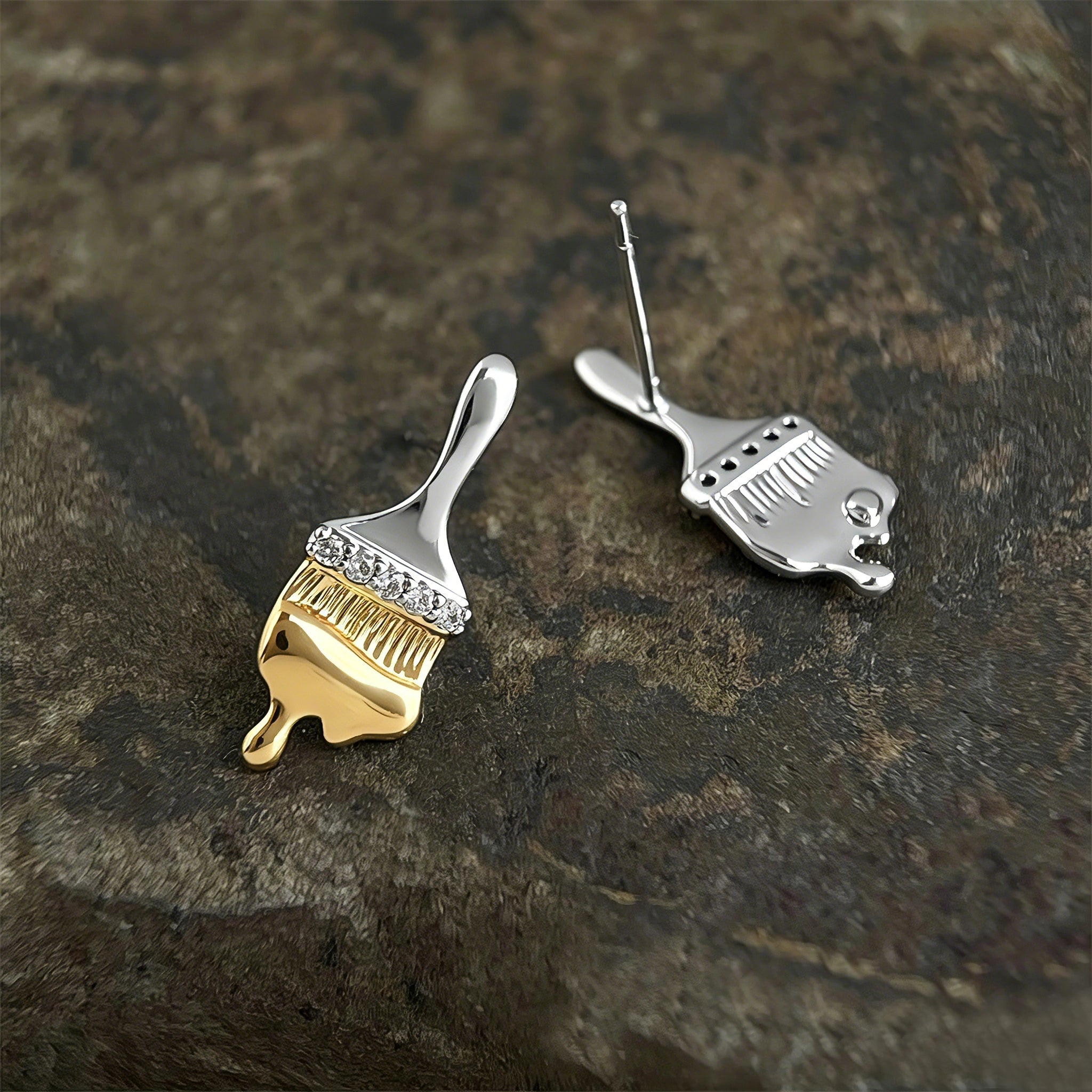 These stud earrings are designed in the shape of a paintbrush, featuring a two-tone finish with a polished silver-toned handle and a gold-toned brush head. The ferrule of the brush is encrusted with sparkling cubic zirconia stones, and the gold-toned bristles are styled to look like dripping, liquid gold paint, creating a vivid, artistic effect. Crafted with a thick 18K real gold plating, they combine a playful, creative design with elegant, eye-catching sparkle.
