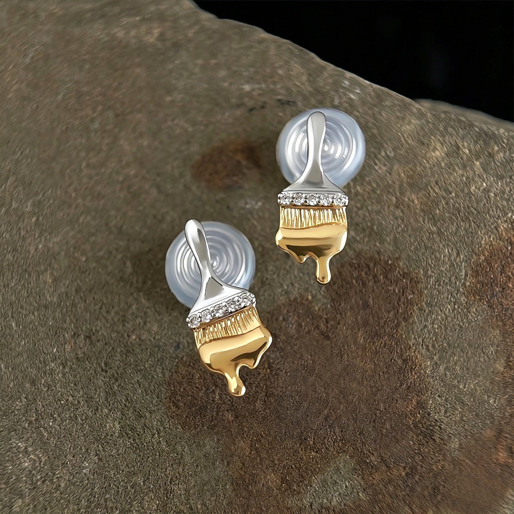 These stud earrings are designed in the shape of a paintbrush, featuring a two-tone finish with a polished silver-toned handle and a gold-toned brush head. The ferrule of the brush is encrusted with sparkling cubic zirconia stones, and the gold-toned bristles are styled to look like dripping, liquid gold paint, creating a vivid, artistic effect. Crafted with a thick 18K real gold plating, they combine a playful, creative design with elegant, eye-catching sparkle.