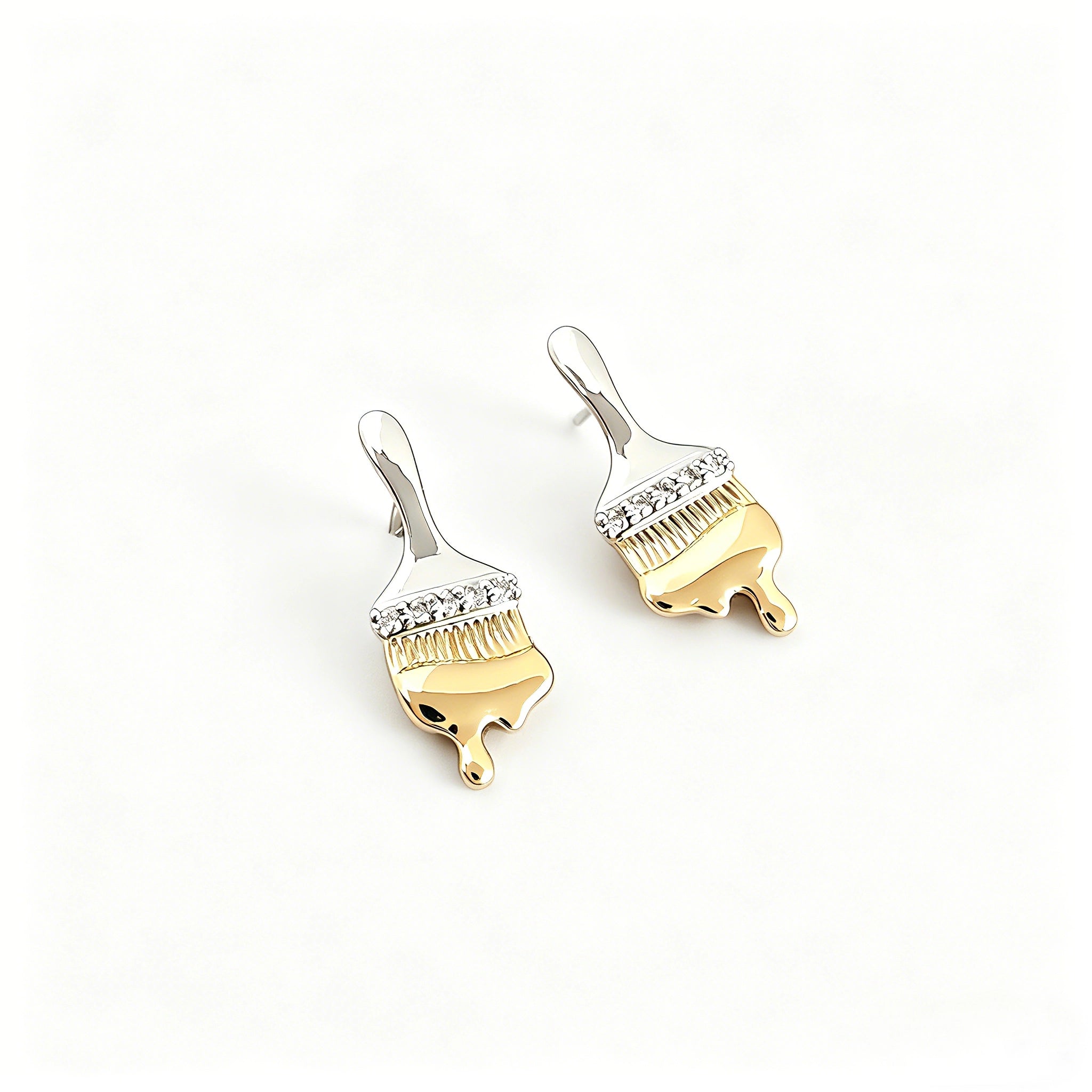 These stud earrings are designed in the shape of a paintbrush, featuring a two-tone finish with a polished silver-toned handle and a gold-toned brush head. The ferrule of the brush is encrusted with sparkling cubic zirconia stones, and the gold-toned bristles are styled to look like dripping, liquid gold paint, creating a vivid, artistic effect. Crafted with a thick 18K real gold plating, they combine a playful, creative design with elegant, eye-catching sparkle.