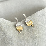 These stud earrings are designed in the shape of a paintbrush, featuring a two-tone finish with a polished silver-toned handle and a gold-toned brush head. The ferrule of the brush is encrusted with sparkling cubic zirconia stones, and the gold-toned bristles are styled to look like dripping, liquid gold paint, creating a vivid, artistic effect. Crafted with a thick 18K real gold plating, they combine a playful, creative design with elegant, eye-catching sparkle.