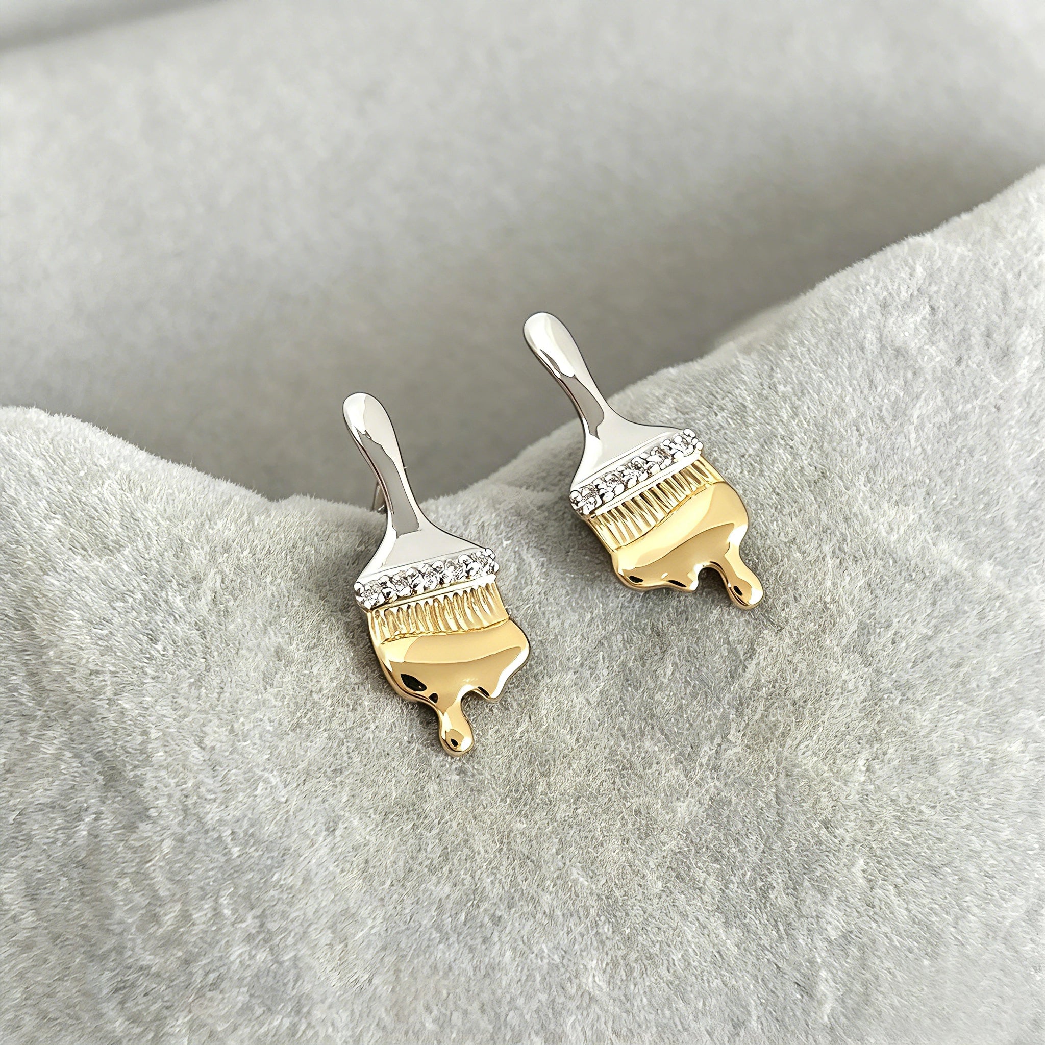These stud earrings are designed in the shape of a paintbrush, featuring a two-tone finish with a polished silver-toned handle and a gold-toned brush head. The ferrule of the brush is encrusted with sparkling cubic zirconia stones, and the gold-toned bristles are styled to look like dripping, liquid gold paint, creating a vivid, artistic effect. Crafted with a thick 18K real gold plating, they combine a playful, creative design with elegant, eye-catching sparkle.