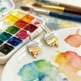 These stud earrings are designed in the shape of a paintbrush, featuring a two-tone finish with a polished silver-toned handle and a gold-toned brush head. The ferrule of the brush is encrusted with sparkling cubic zirconia stones, and the gold-toned bristles are styled to look like dripping, liquid gold paint, creating a vivid, artistic effect. Crafted with a thick 18K real gold plating, they combine a playful, creative design with elegant, eye-catching sparkle.