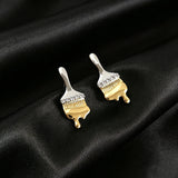 These stud earrings are designed in the shape of a paintbrush, featuring a two-tone finish with a polished silver-toned handle and a gold-toned brush head. The ferrule of the brush is encrusted with sparkling cubic zirconia stones, and the gold-toned bristles are styled to look like dripping, liquid gold paint, creating a vivid, artistic effect. Crafted with a thick 18K real gold plating, they combine a playful, creative design with elegant, eye-catching sparkle.