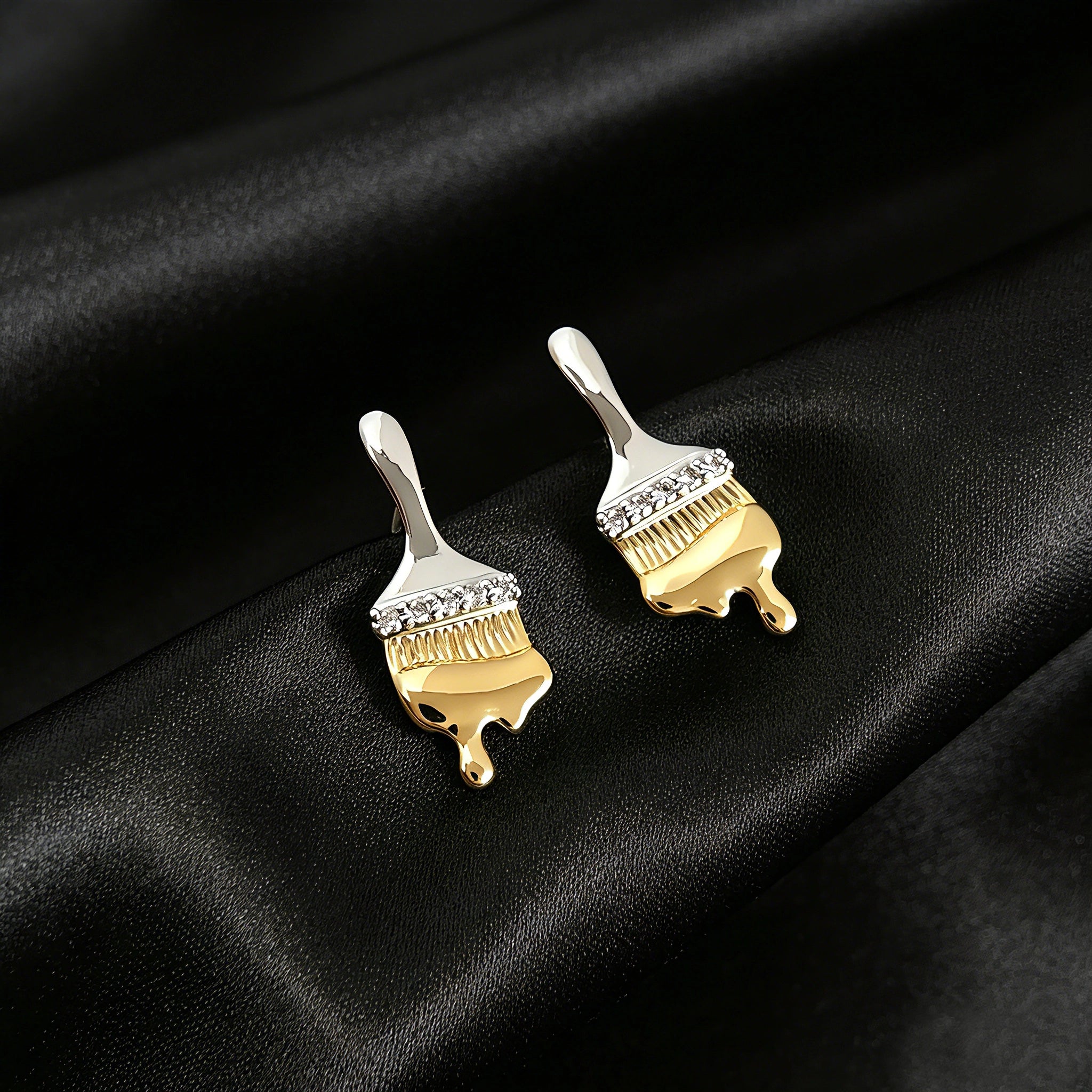 These stud earrings are designed in the shape of a paintbrush, featuring a two-tone finish with a polished silver-toned handle and a gold-toned brush head. The ferrule of the brush is encrusted with sparkling cubic zirconia stones, and the gold-toned bristles are styled to look like dripping, liquid gold paint, creating a vivid, artistic effect. Crafted with a thick 18K real gold plating, they combine a playful, creative design with elegant, eye-catching sparkle.