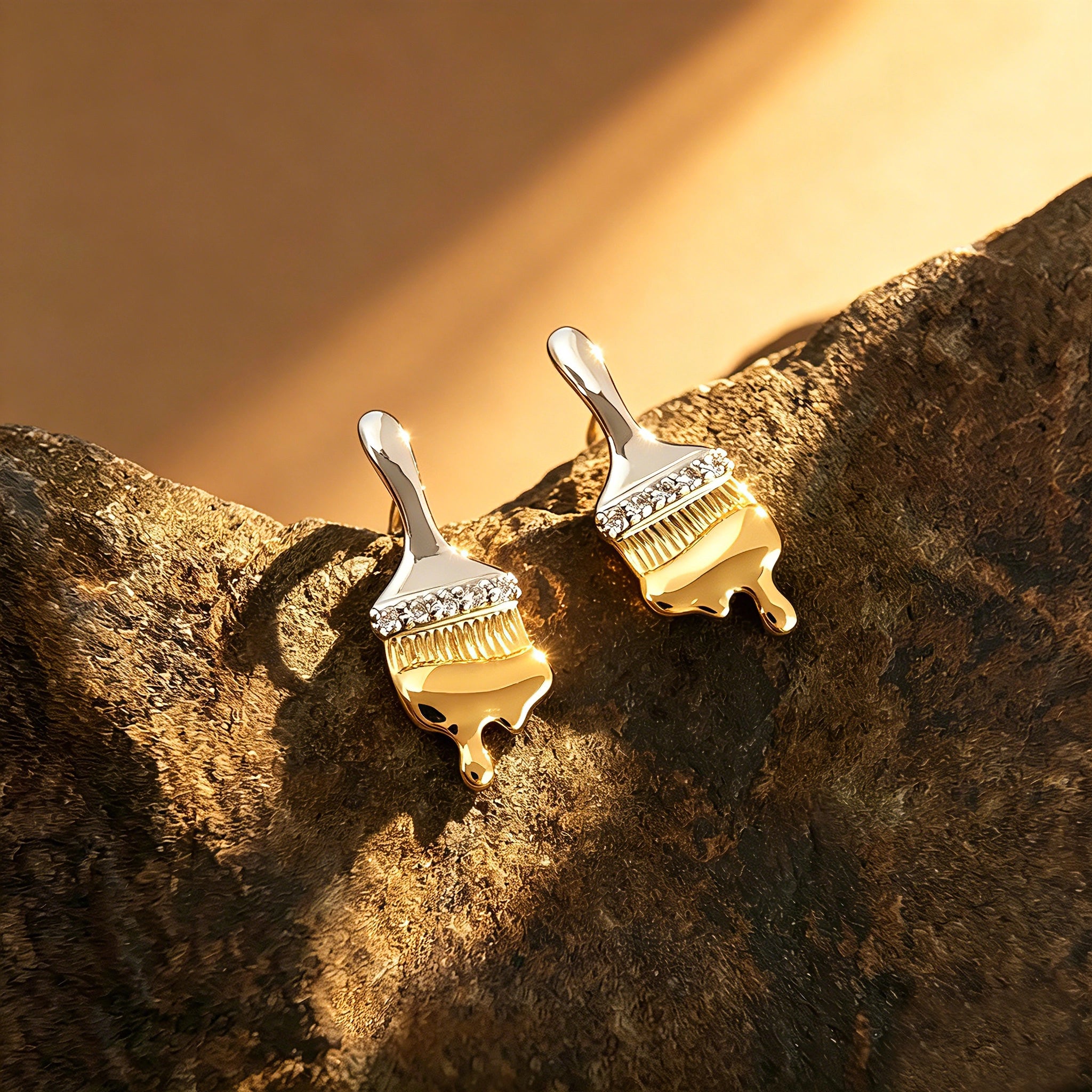 These stud earrings are designed in the shape of a paintbrush, featuring a two-tone finish with a polished silver-toned handle and a gold-toned brush head. The ferrule of the brush is encrusted with sparkling cubic zirconia stones, and the gold-toned bristles are styled to look like dripping, liquid gold paint, creating a vivid, artistic effect. Crafted with a thick 18K real gold plating, they combine a playful, creative design with elegant, eye-catching sparkle.