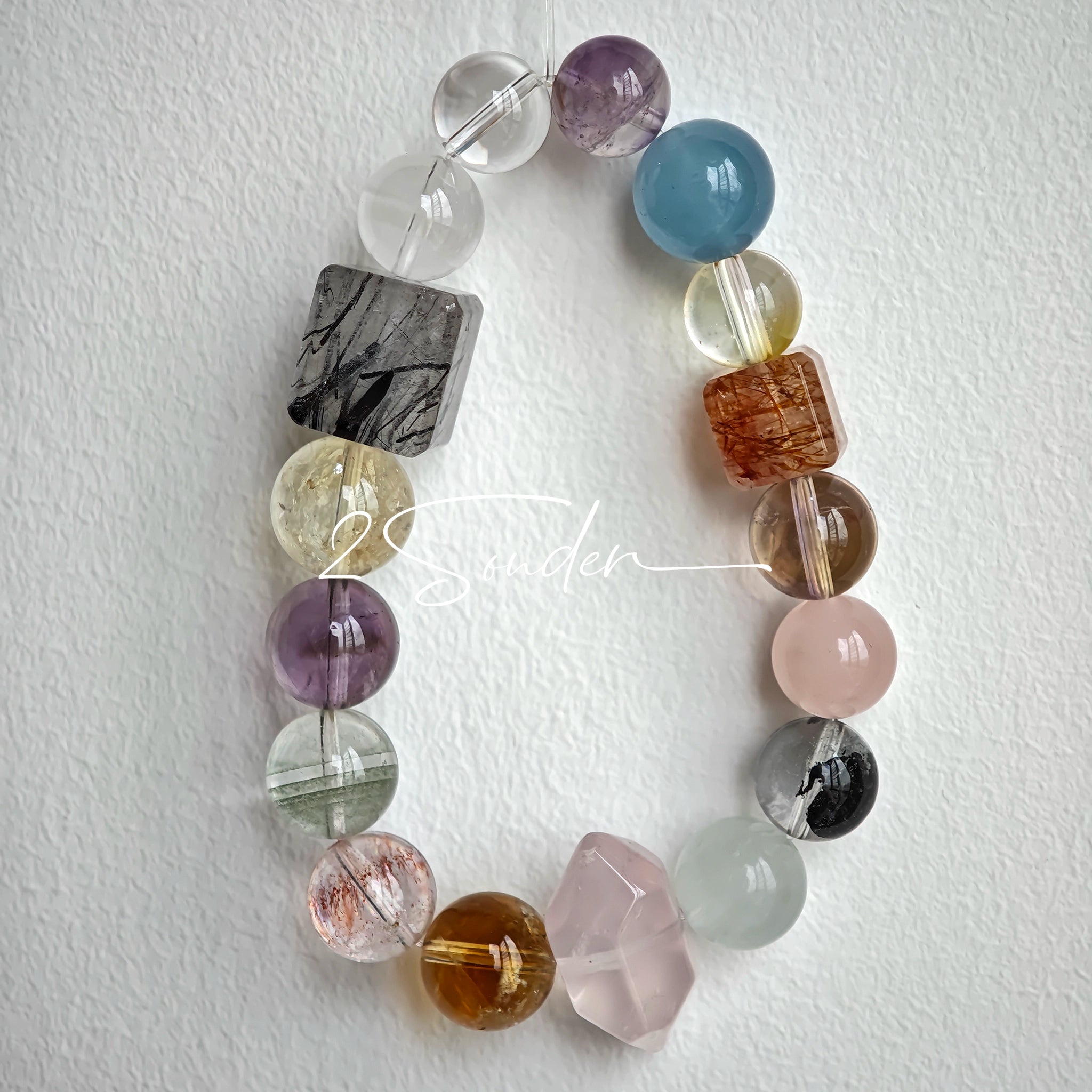 This stunning multi-gem crystal bracelet is a vibrant rainbow of natural stones, featuring smooth round beads in hues of clear, amethyst, sky blue, soft pink, golden, and smoky brown, interspersed with striking square and geometric-cut accents.
Each bead showcases unique inclusions: delicate black rutile in the gray square, fiery red hair-like strands in the brown square, and subtle mineral veining throughout, creating a one-of-a-kind, textured masterpiece.
The eclectic mix of gemstones—from amethyst and ro