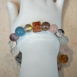 This stunning multi-gem crystal bracelet is a vibrant rainbow of natural stones, featuring smooth round beads in hues of clear, amethyst, sky blue, soft pink, golden, and smoky brown, interspersed with striking square and geometric-cut accents.
Each bead showcases unique inclusions: delicate black rutile in the gray square, fiery red hair-like strands in the brown square, and subtle mineral veining throughout, creating a one-of-a-kind, textured masterpiece.
The eclectic mix of gemstones—from amethyst and ro