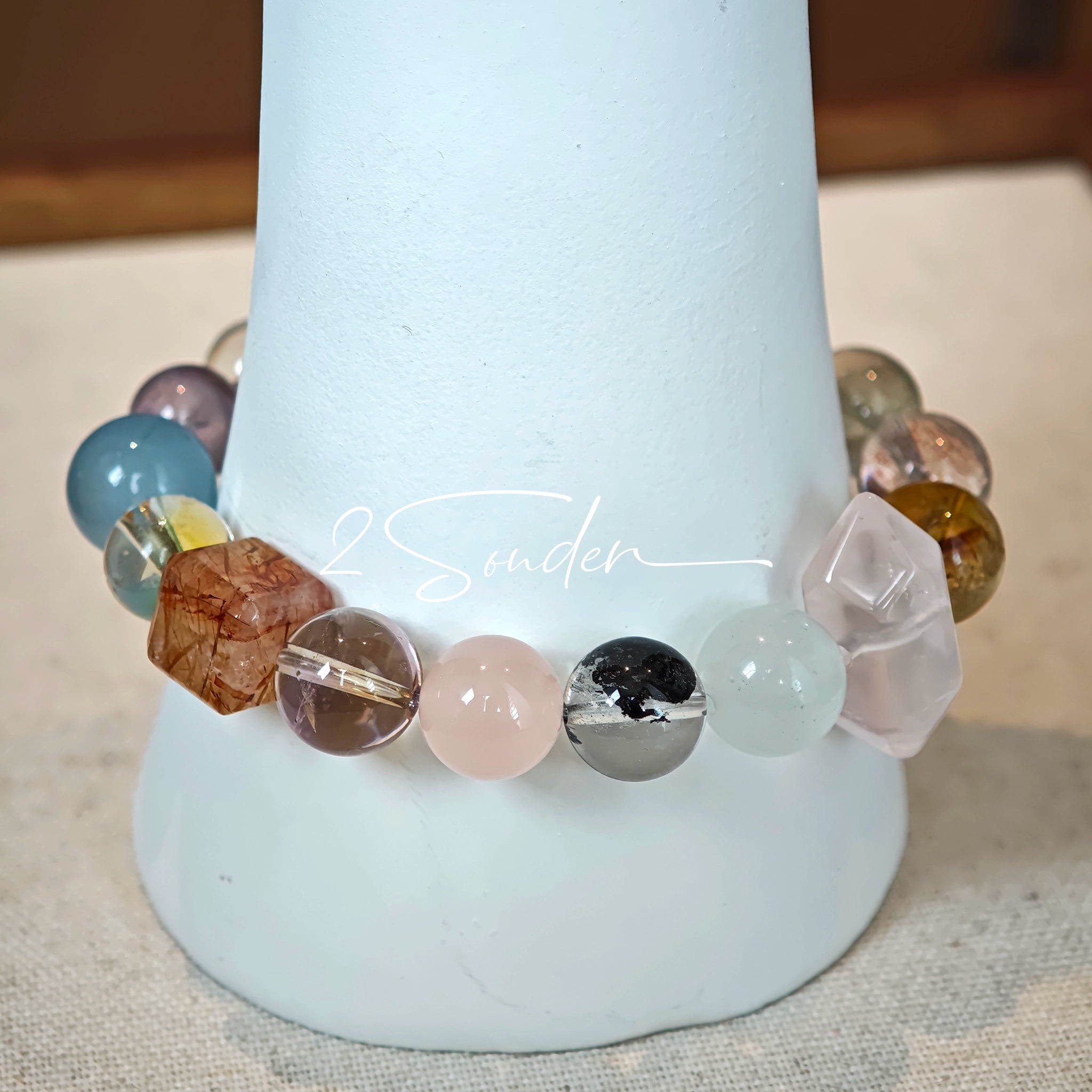 This stunning multi-gem crystal bracelet is a vibrant rainbow of natural stones, featuring smooth round beads in hues of clear, amethyst, sky blue, soft pink, golden, and smoky brown, interspersed with striking square and geometric-cut accents.
Each bead showcases unique inclusions: delicate black rutile in the gray square, fiery red hair-like strands in the brown square, and subtle mineral veining throughout, creating a one-of-a-kind, textured masterpiece.
The eclectic mix of gemstones—from amethyst and ro