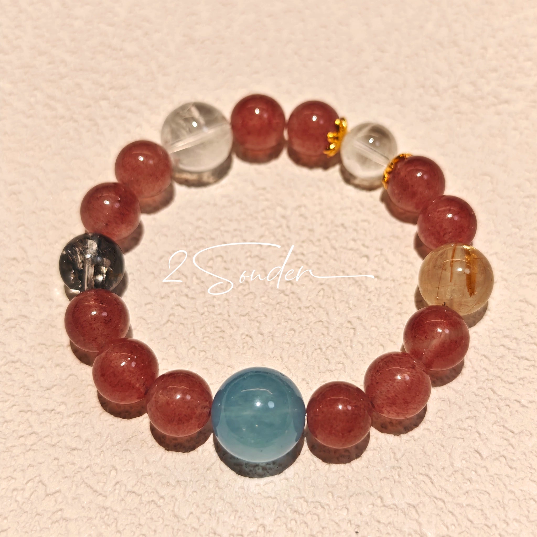 This bracelet is centered around a prominent round light blue aquamarine bead, surrounded by rich reddish-brown strawberry quartz beads. It is accented with clear crystal, black rutilated quartz, and golden rutilated quartz beads, along with delicate gold spacers, creating a harmonious natural stone design.