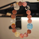 This bracelet is centered around a prominent round light blue aquamarine bead, surrounded by rich reddish-brown strawberry quartz beads. It is accented with clear crystal, black rutilated quartz, and golden rutilated quartz beads, along with delicate gold spacers, creating a harmonious natural stone design.