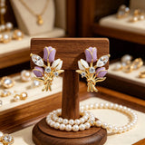 These stud earrings feature an elegant bouquet design, crafted with a thick 18K real gold plating. The arrangement showcases delicate purple and white enamel tulip flowers, accented with sparkling marquise and round-cut cubic zirconia stones, all set in a polished gold-tone metal structure tied with a decorative knot at the base. The overall style is romantic and refined, perfect for adding a touch of floral charm to any look.