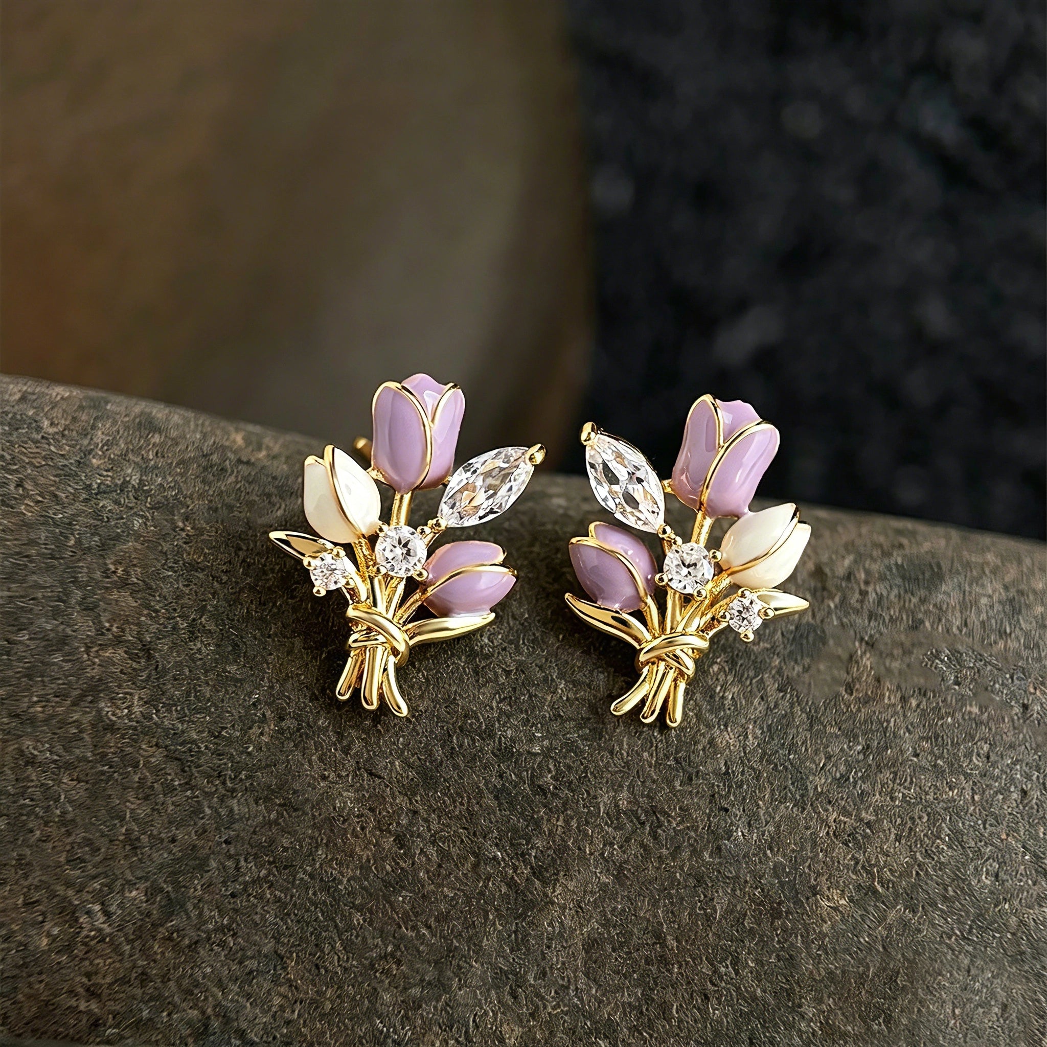 These stud earrings feature an elegant bouquet design, crafted with a thick 18K real gold plating. The arrangement showcases delicate purple and white enamel tulip flowers, accented with sparkling marquise and round-cut cubic zirconia stones, all set in a polished gold-tone metal structure tied with a decorative knot at the base. The overall style is romantic and refined, perfect for adding a touch of floral charm to any look.