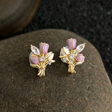 These stud earrings feature an elegant bouquet design, crafted with a thick 18K real gold plating. The arrangement showcases delicate purple and white enamel tulip flowers, accented with sparkling marquise and round-cut cubic zirconia stones, all set in a polished gold-tone metal structure tied with a decorative knot at the base. The overall style is romantic and refined, perfect for adding a touch of floral charm to any look.