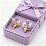 These stud earrings feature an elegant bouquet design, crafted with a thick 18K real gold plating. The arrangement showcases delicate purple and white enamel tulip flowers, accented with sparkling marquise and round-cut cubic zirconia stones, all set in a polished gold-tone metal structure tied with a decorative knot at the base. The overall style is romantic and refined, perfect for adding a touch of floral charm to any look.
