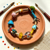 This handmade boho bracelet bursts with vibrant dopamine colors, featuring hand-painted clay beads, twisted linen threads, and quirky sculpted charms. Every bead and wrapped fiber is carefully crafted, bringing playful, retro artistic vibes to your wrist. It is a unique wearable art piece full of whimsical details.