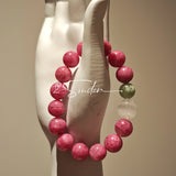 This beaded bracelet is mainly made of round, rosy-pink rhodonite beads with a delicate marbled texture. It is accented by one pale green Xiuyan jade bead (in the "green grape milk cap" style) and one translucent white phantom quartz bead, creating a soft color contrast against the dominant pink hue. The smooth, polished stones give the bracelet a gentle, natural look.