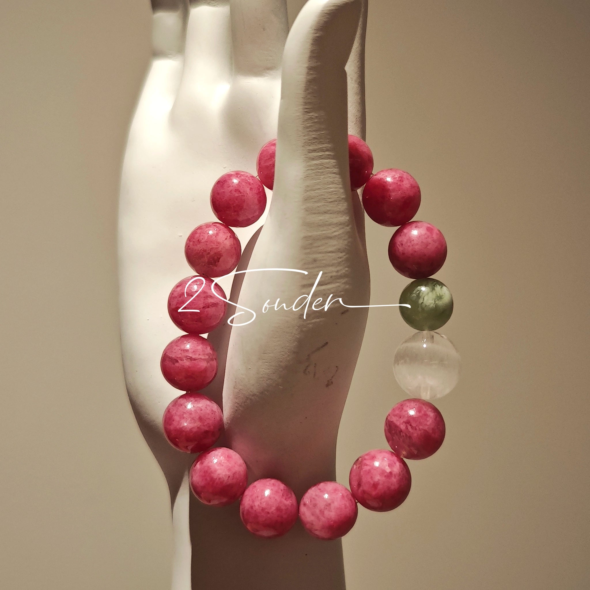 This beaded bracelet is mainly made of round, rosy-pink rhodonite beads with a delicate marbled texture. It is accented by one pale green Xiuyan jade bead (in the "green grape milk cap" style) and one translucent white phantom quartz bead, creating a soft color contrast against the dominant pink hue. The smooth, polished stones give the bracelet a gentle, natural look.