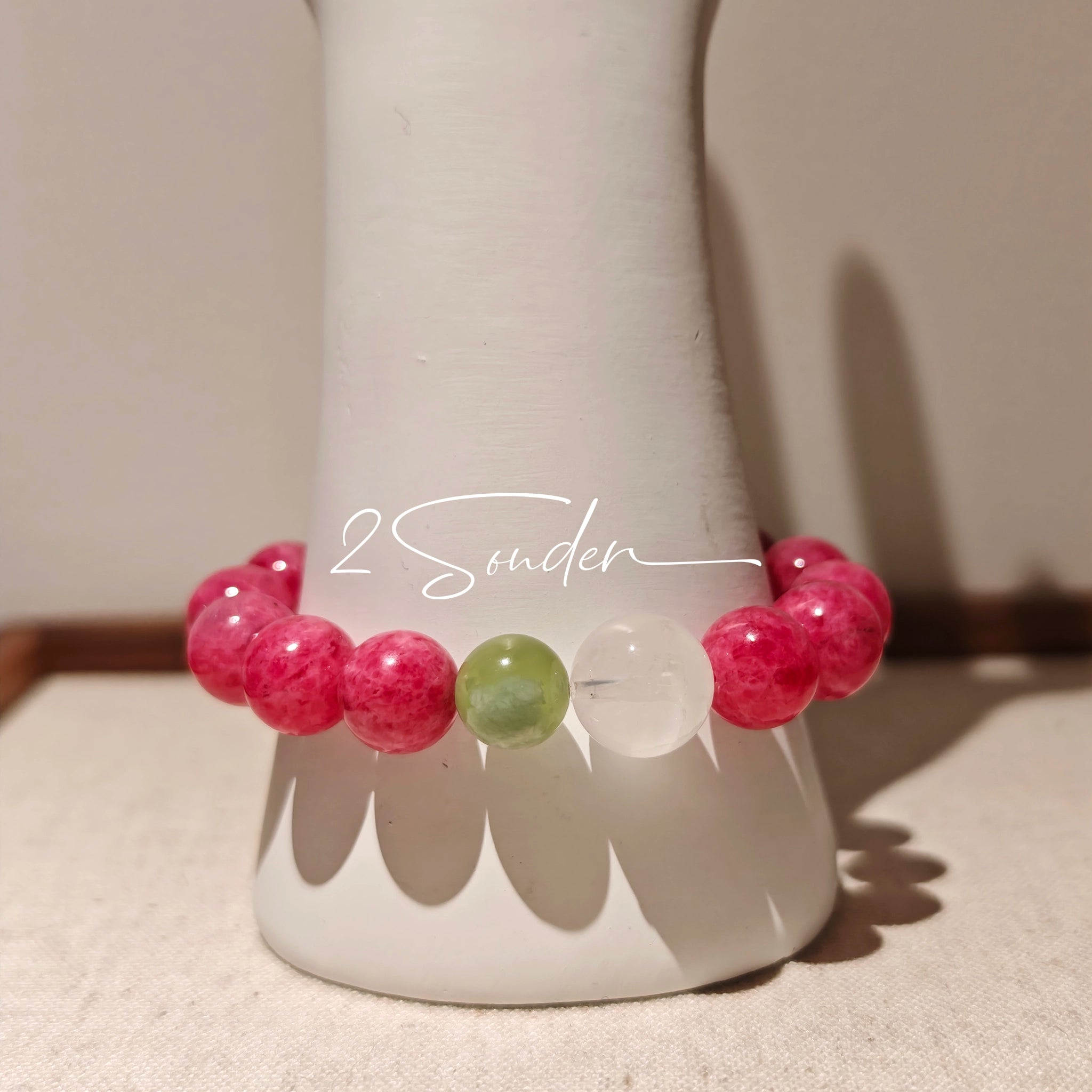 This beaded bracelet is mainly made of round, rosy-pink rhodonite beads with a delicate marbled texture. It is accented by one pale green Xiuyan jade bead (in the "green grape milk cap" style) and one translucent white phantom quartz bead, creating a soft color contrast against the dominant pink hue. The smooth, polished stones give the bracelet a gentle, natural look.