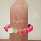 This beaded bracelet is mainly made of round, rosy-pink rhodonite beads with a delicate marbled texture. It is accented by one pale green Xiuyan jade bead (in the "green grape milk cap" style) and one translucent white phantom quartz bead, creating a soft color contrast against the dominant pink hue. The smooth, polished stones give the bracelet a gentle, natural look.