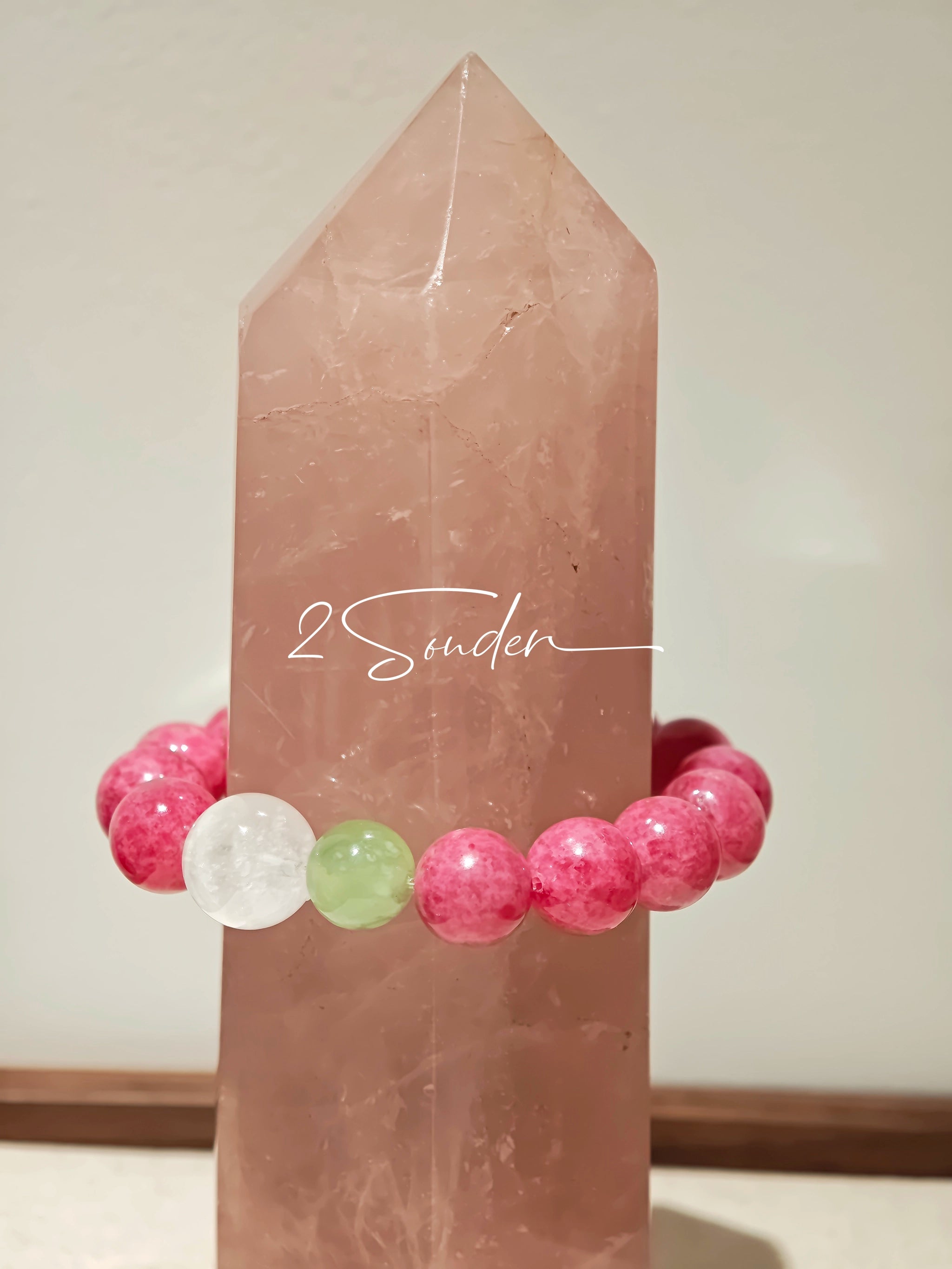 This beaded bracelet is mainly made of round, rosy-pink rhodonite beads with a delicate marbled texture. It is accented by one pale green Xiuyan jade bead (in the "green grape milk cap" style) and one translucent white phantom quartz bead, creating a soft color contrast against the dominant pink hue. The smooth, polished stones give the bracelet a gentle, natural look.