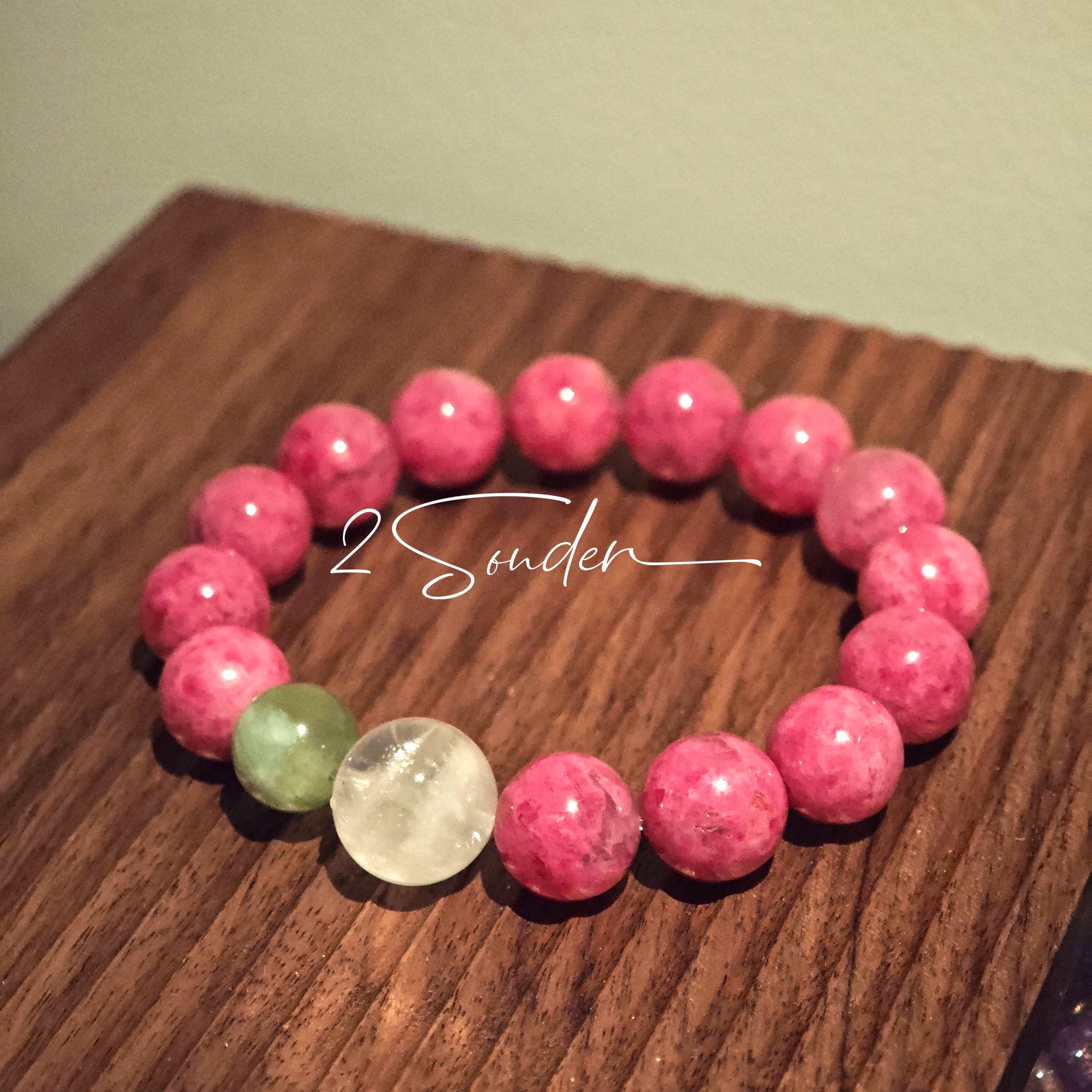 This beaded bracelet is mainly made of round, rosy-pink rhodonite beads with a delicate marbled texture. It is accented by one pale green Xiuyan jade bead (in the "green grape milk cap" style) and one translucent white phantom quartz bead, creating a soft color contrast against the dominant pink hue. The smooth, polished stones give the bracelet a gentle, natural look.
