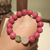 This beaded bracelet is mainly made of round, rosy-pink rhodonite beads with a delicate marbled texture. It is accented by one pale green Xiuyan jade bead (in the "green grape milk cap" style) and one translucent white phantom quartz bead, creating a soft color contrast against the dominant pink hue. The smooth, polished stones give the bracelet a gentle, natural look.