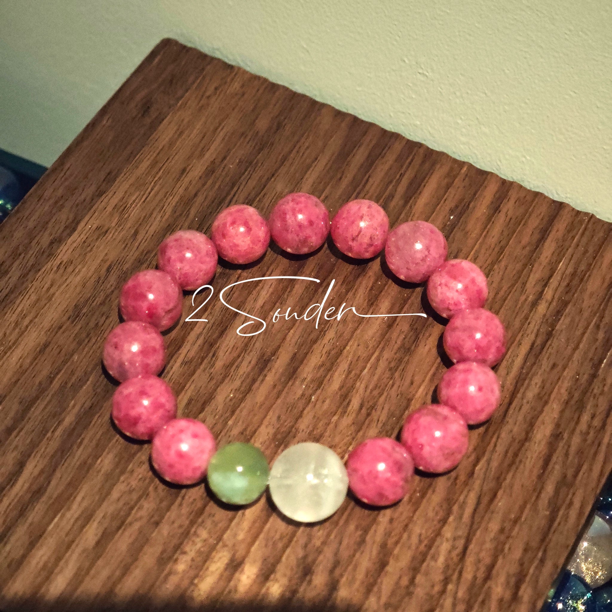 This beaded bracelet is mainly made of round, rosy-pink rhodonite beads with a delicate marbled texture. It is accented by one pale green Xiuyan jade bead (in the "green grape milk cap" style) and one translucent white phantom quartz bead, creating a soft color contrast against the dominant pink hue. The smooth, polished stones give the bracelet a gentle, natural look.