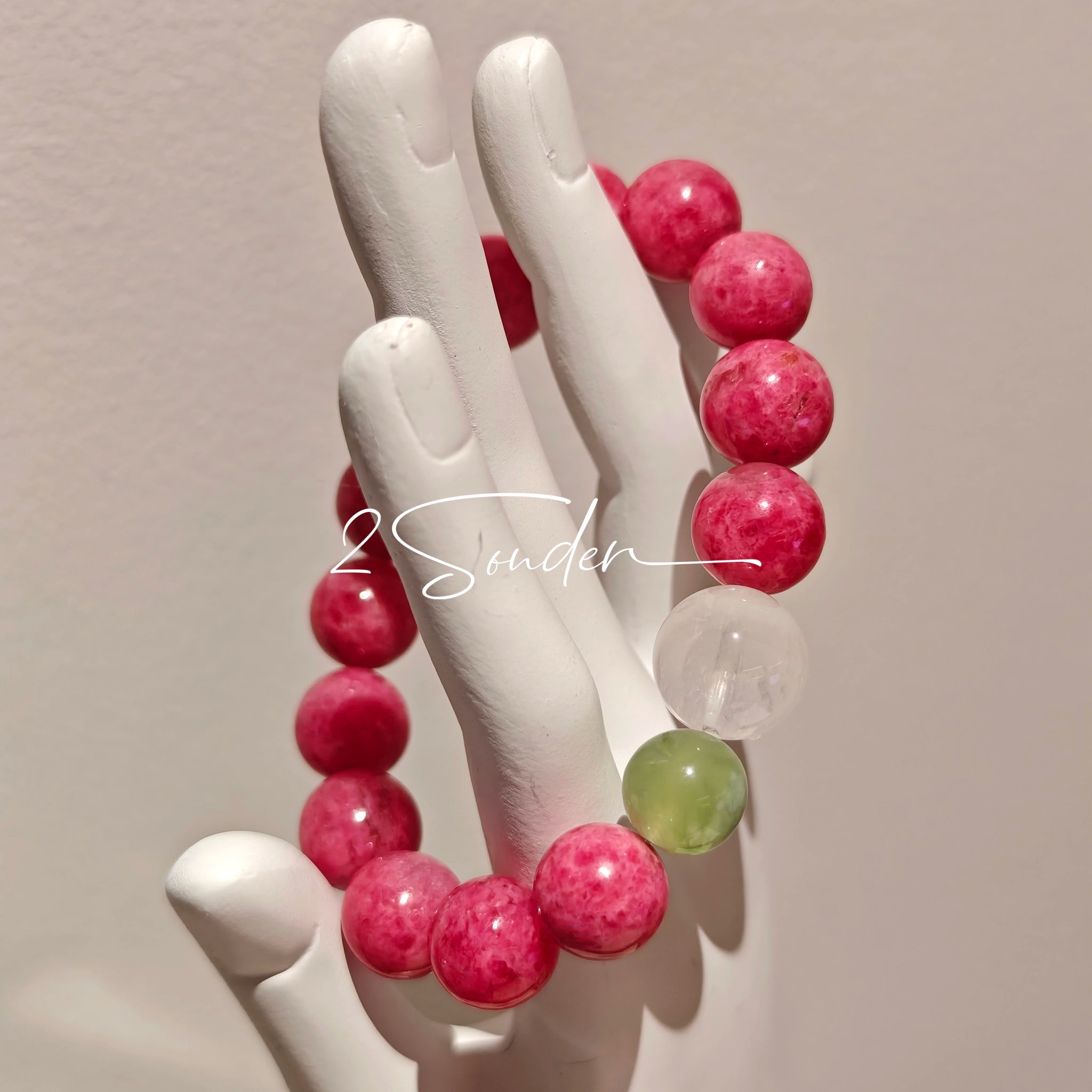 This beaded bracelet is mainly made of round, rosy-pink rhodonite beads with a delicate marbled texture. It is accented by one pale green Xiuyan jade bead (in the "green grape milk cap" style) and one translucent white phantom quartz bead, creating a soft color contrast against the dominant pink hue. The smooth, polished stones give the bracelet a gentle, natural look.