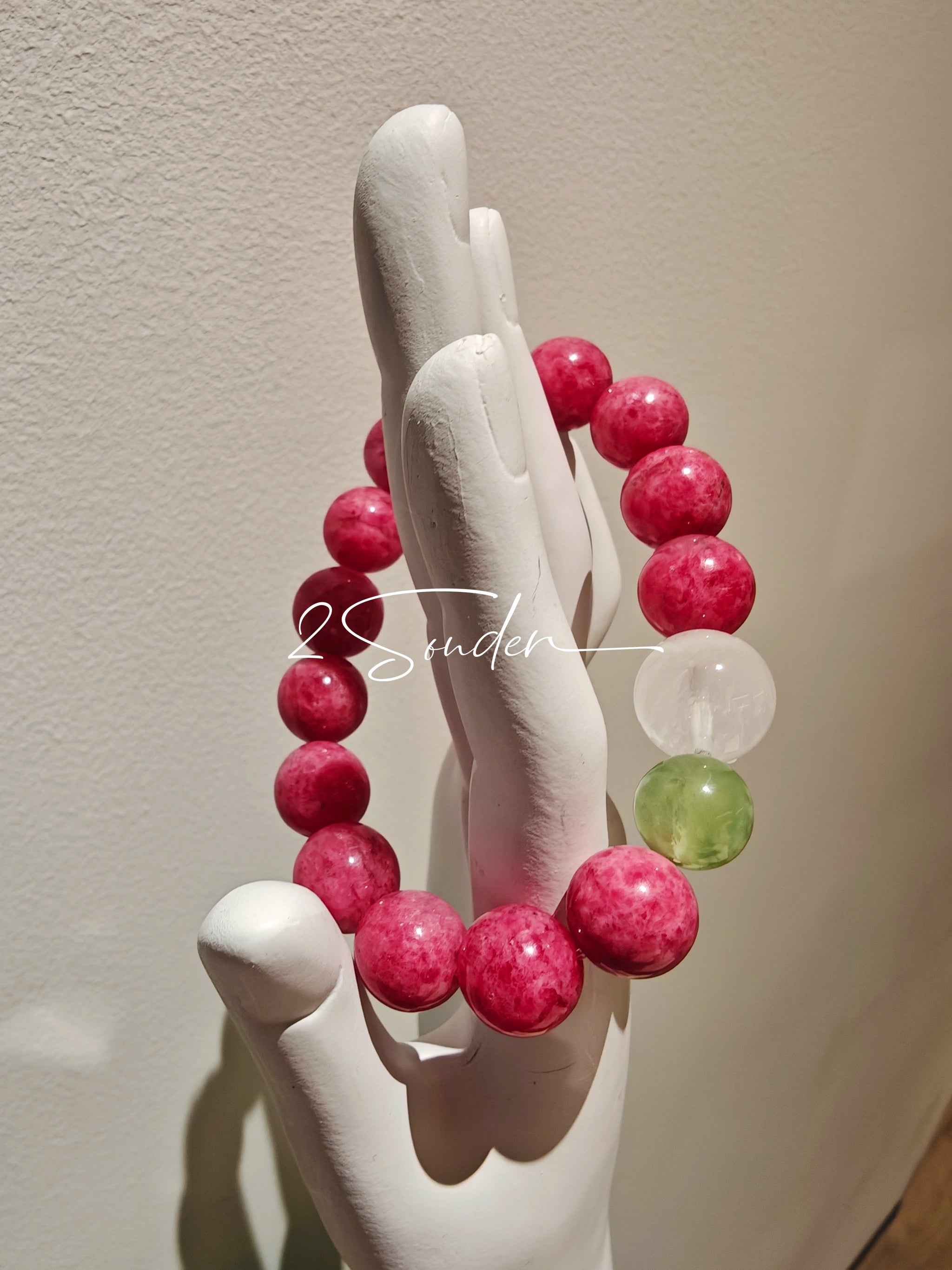 This beaded bracelet is mainly made of round, rosy-pink rhodonite beads with a delicate marbled texture. It is accented by one pale green Xiuyan jade bead (in the "green grape milk cap" style) and one translucent white phantom quartz bead, creating a soft color contrast against the dominant pink hue. The smooth, polished stones give the bracelet a gentle, natural look.
