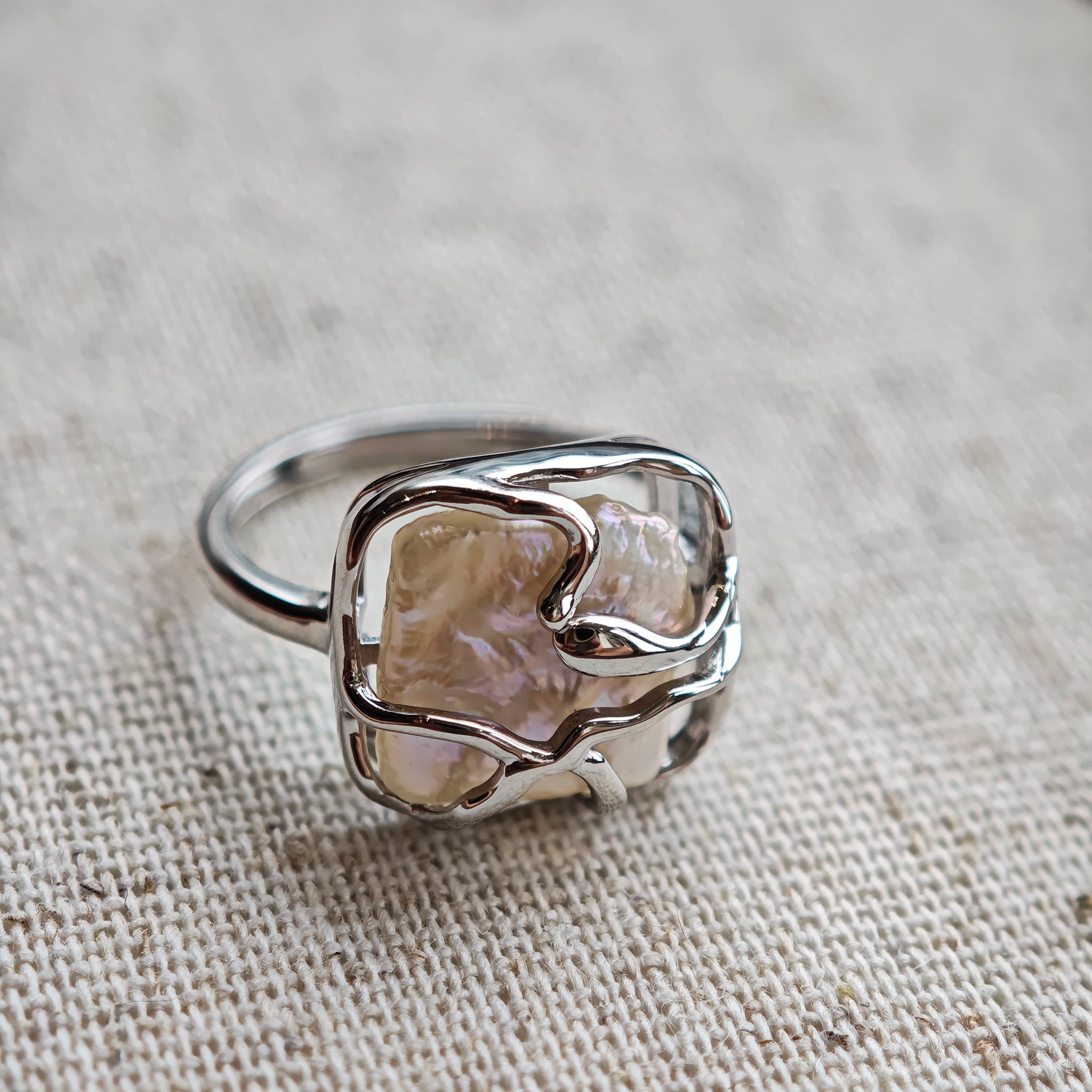 This adjustable S925 sterling silver ring features a striking baroque pearl at its center, set within an organic, flowing silver frame that wraps around the irregular, lustrous gemstone. The open, sculptural metalwork creates a modern, artistic silhouette, showcasing the pearl's natural, one-of-a-kind texture and soft iridescence. The sleek, polished silver band complements the unique pearl, making it a standout piece with a contemporary, bohemian aesthetic.