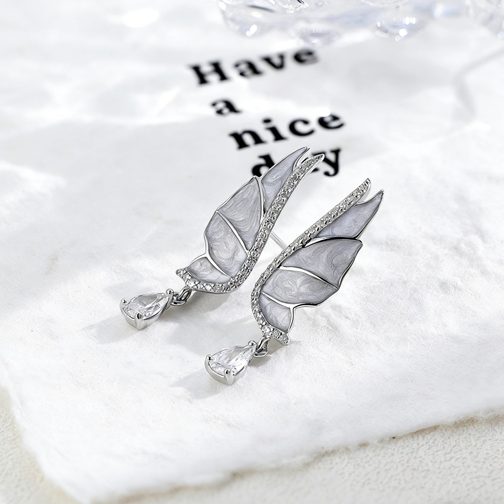 These S925 sterling silver earrings are designed in the shape of delicate butterfly wings, with a resin craft on the wing surfaces and a row of shimmering cubic zirconia along the edges. A teardrop-cut clear gemstone dangles elegantly from the bottom of each wing, adding a graceful and sparkling touch to the overall design.
